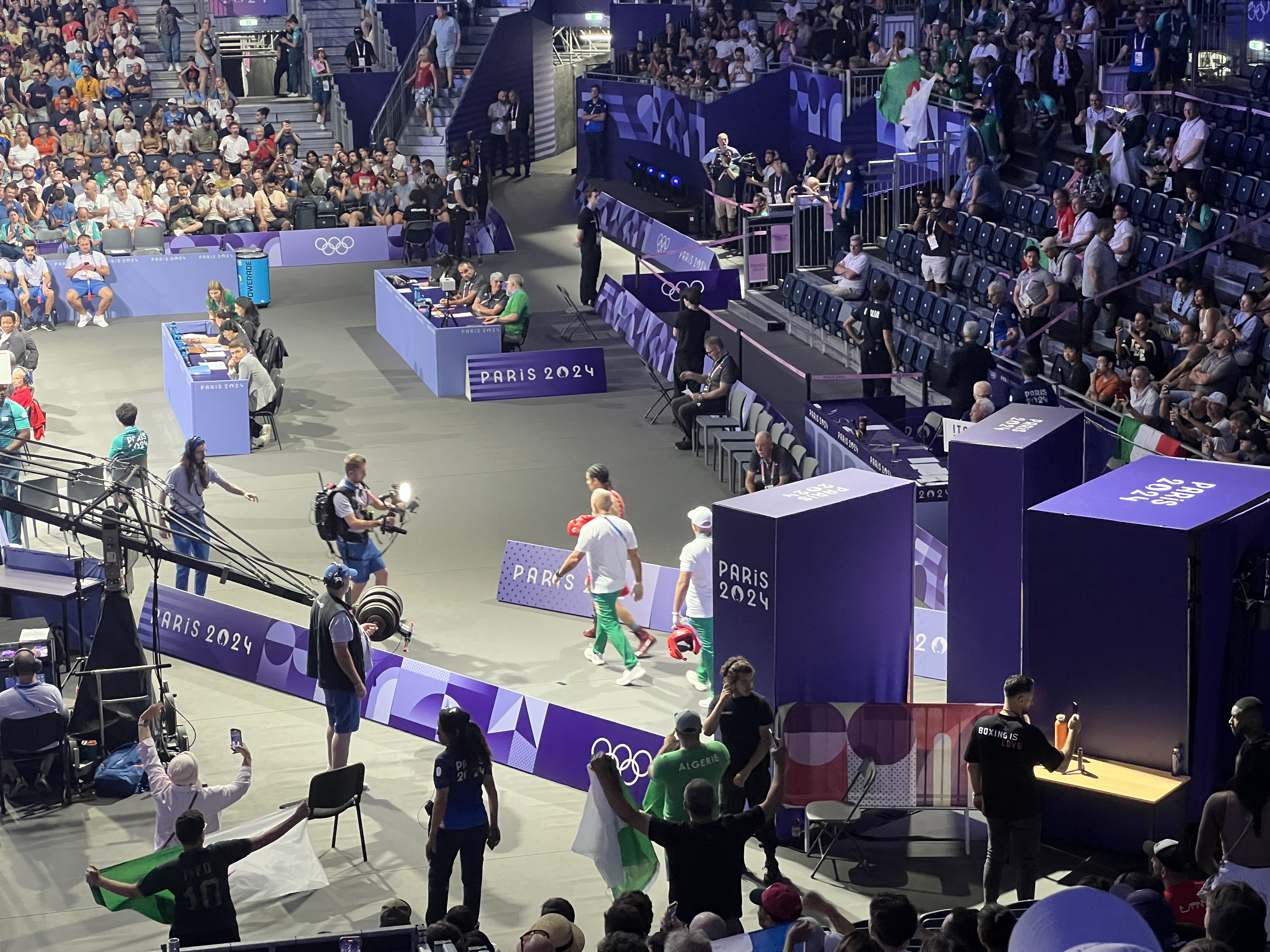 Interior de un pabellón deportivo durante los Juegos Olímpicos de París 2024, mostrando atletas compitiendo en una cancha rodeada de público en las gradas, con banderas olímpicas y carteles de "PARIS 2024" visibles.