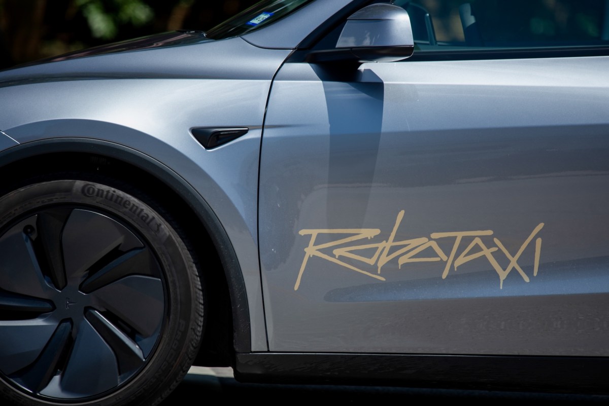 A gray Tesla Cybertaxi with "REBATAX" written in gold lettering on its side, showing the vehicle's wheel and door against a dark background.