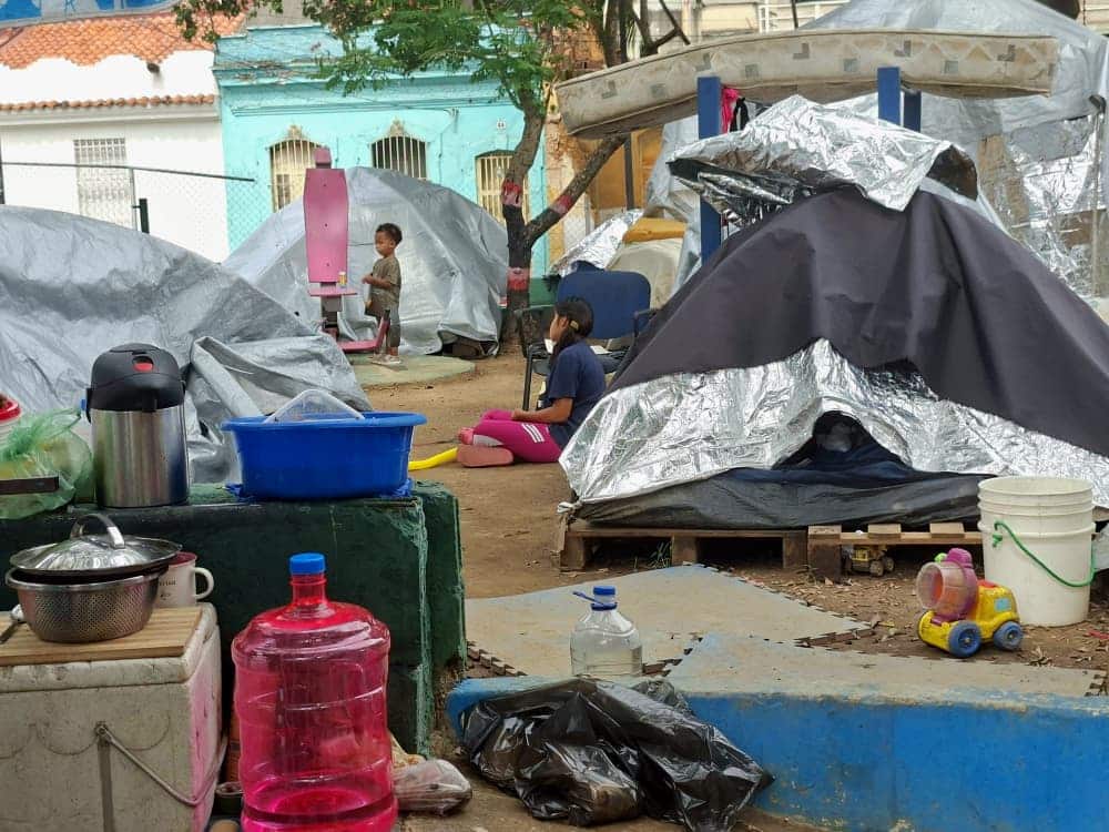 Un campamento improvisado en un terreno con tiendas de campaña plateadas, lonas improvisadas, utensilios de cocina y personas entre estructuras temporales de refugio.