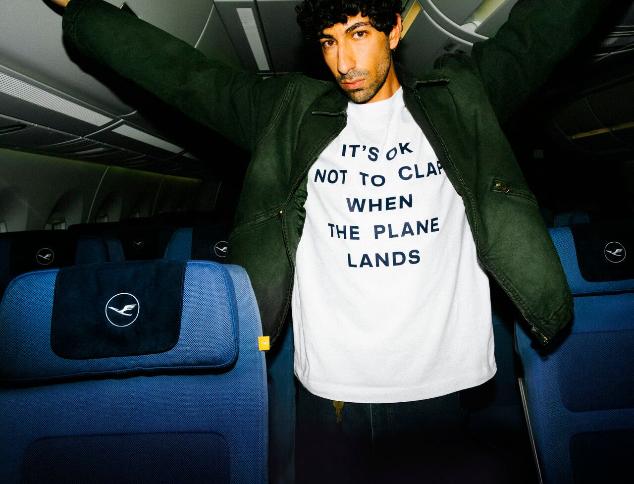 A man wearing a white t-shirt with text and a green jacket sits inside an airplane cabin with blue seats branded with Lufthansa logos.