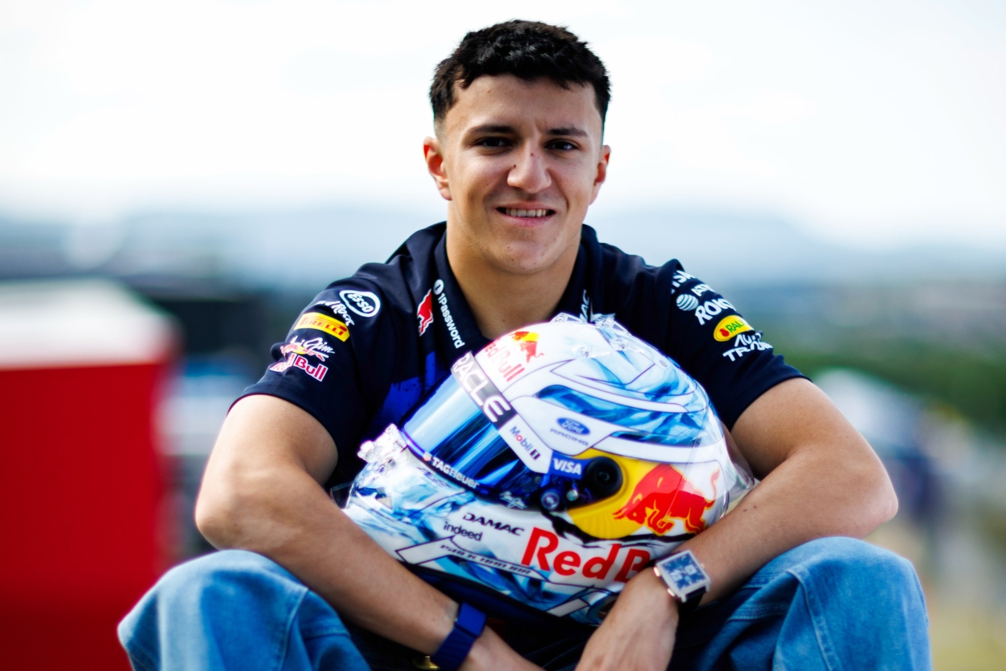 A young racing driver in a navy team uniform with Red Bull logos and sponsor badges holds a white, blue and red racing helmet while smiling at the camera.