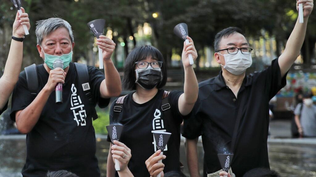 Tres personas con máscaras protectoras sostienen velas encendidas y carteles con caracteres chinos durante una vigilia nocturna en un parque.