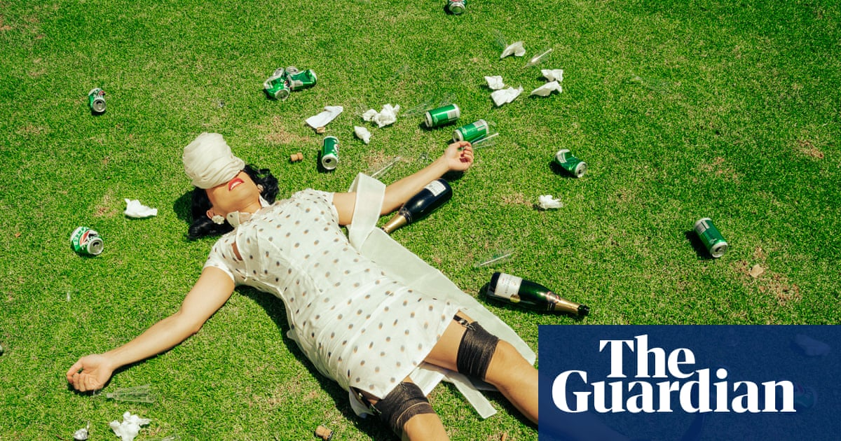 A person lying on grass wearing a white polka-dot dress and bandages, surrounded by scattered beer cans and bottles.