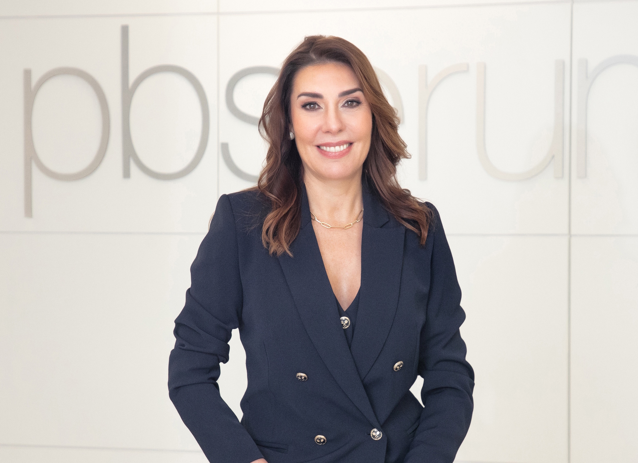 Una mujer profesional con cabello castaño ondulado viste un blazer azul marino de doble botonadura frente a un fondo blanco con letras desenfocadas.