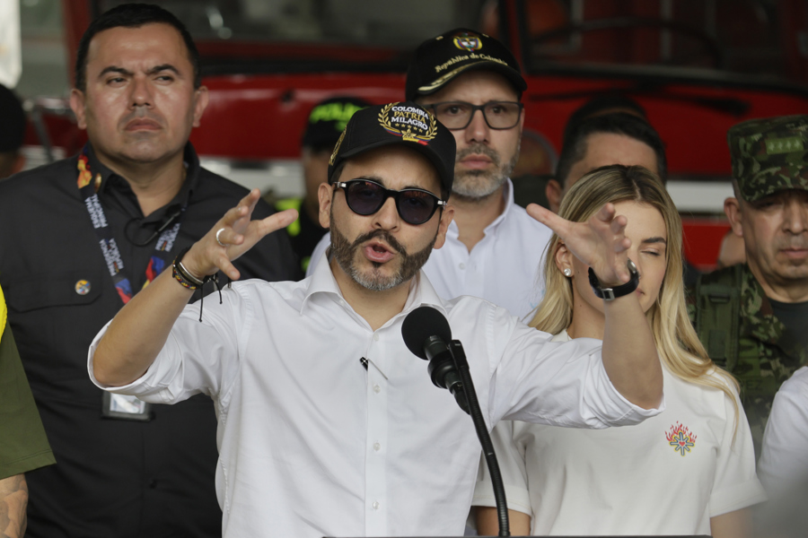 A man wearing sunglasses and a white cap speaks into a microphone, surrounded by uniformed personnel and officials in an emergency zone.