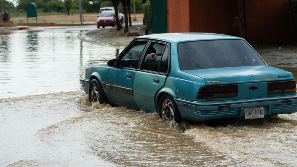 Un automóvil azul turquesa está atrapado en una calle inundada con agua de color marrón que cubre las ruedas del vehículo.