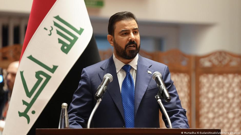 Un hombre viste un traje azul marino y corbata azul, hablando ante dos micrófonos con la bandera de Irak de fondo.