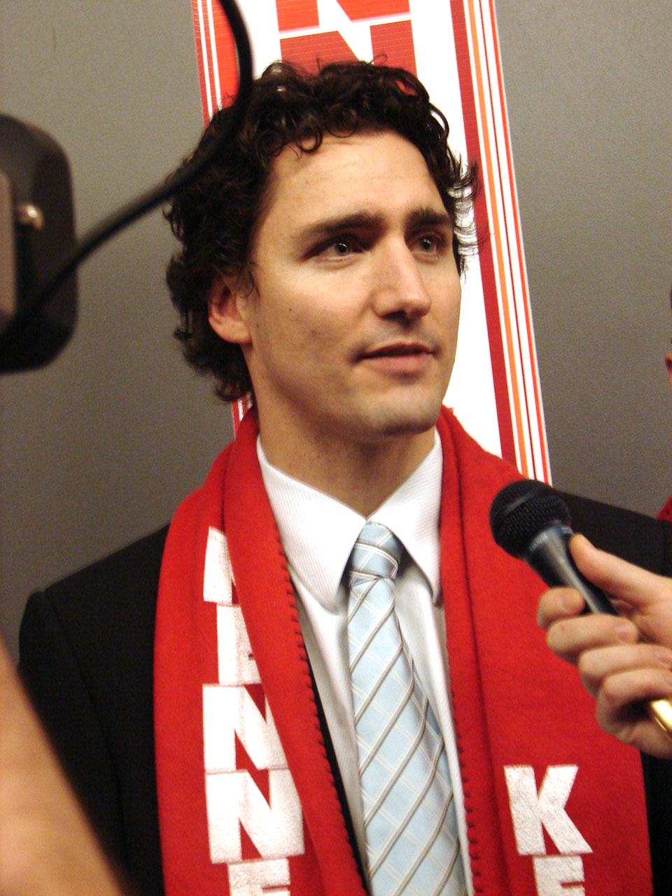A man in a black blazer, white shirt, and striped tie holds a microphone while wearing a red and white scarf with text on it.