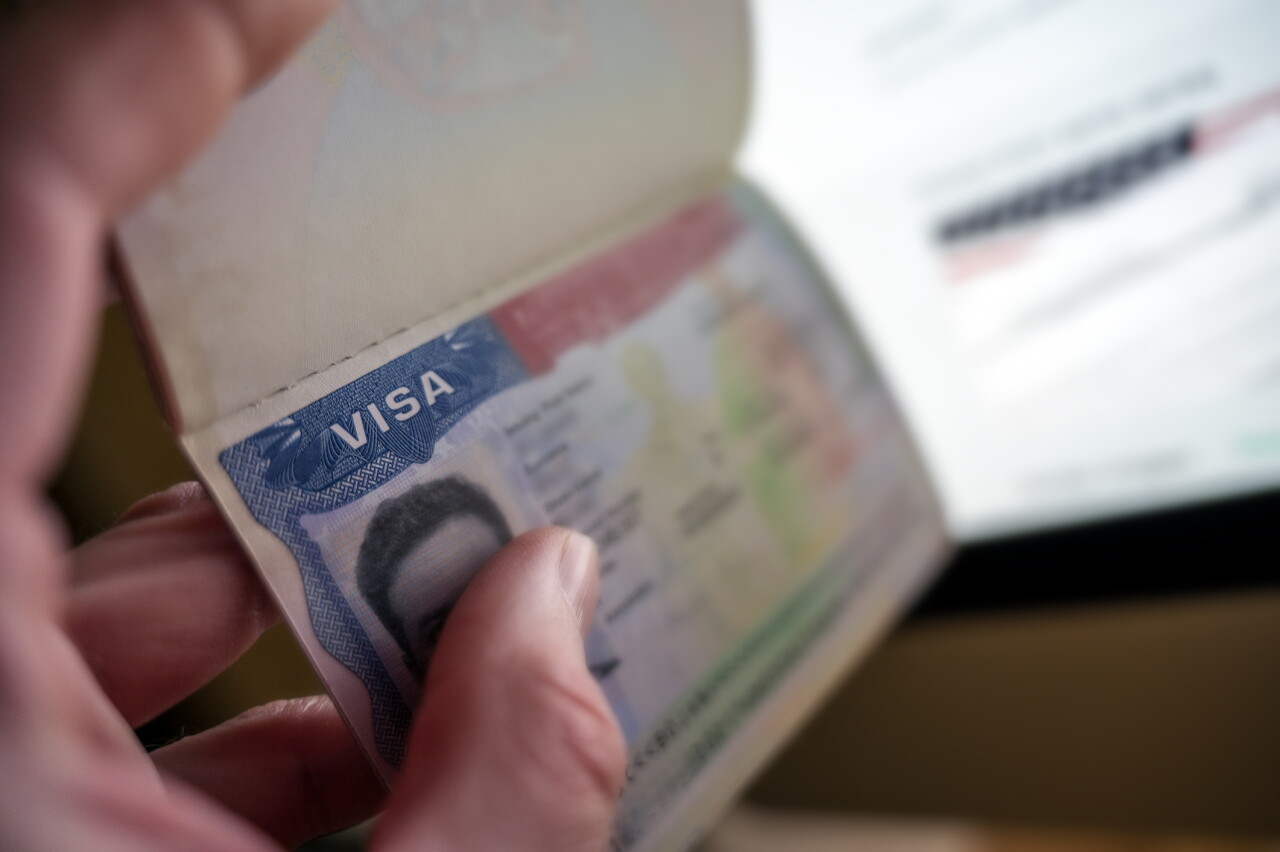 A hand holding an open U.S. passport showing the visa page with a photo visible.