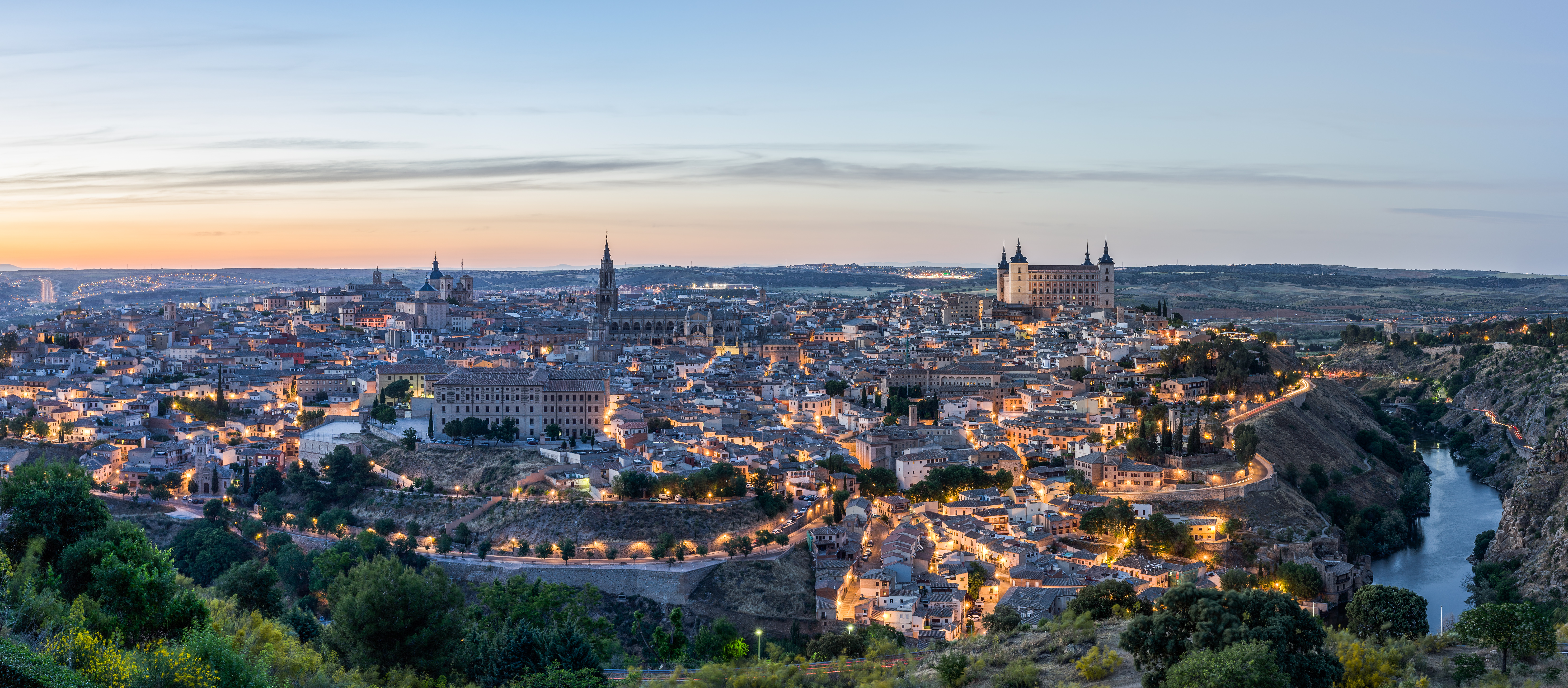 Panoramic view of a medieval city at dusk with a castle of towers on the right hill, illuminated by the golden light of nightfall, surrounded by historic buildings and a river winding around the city.