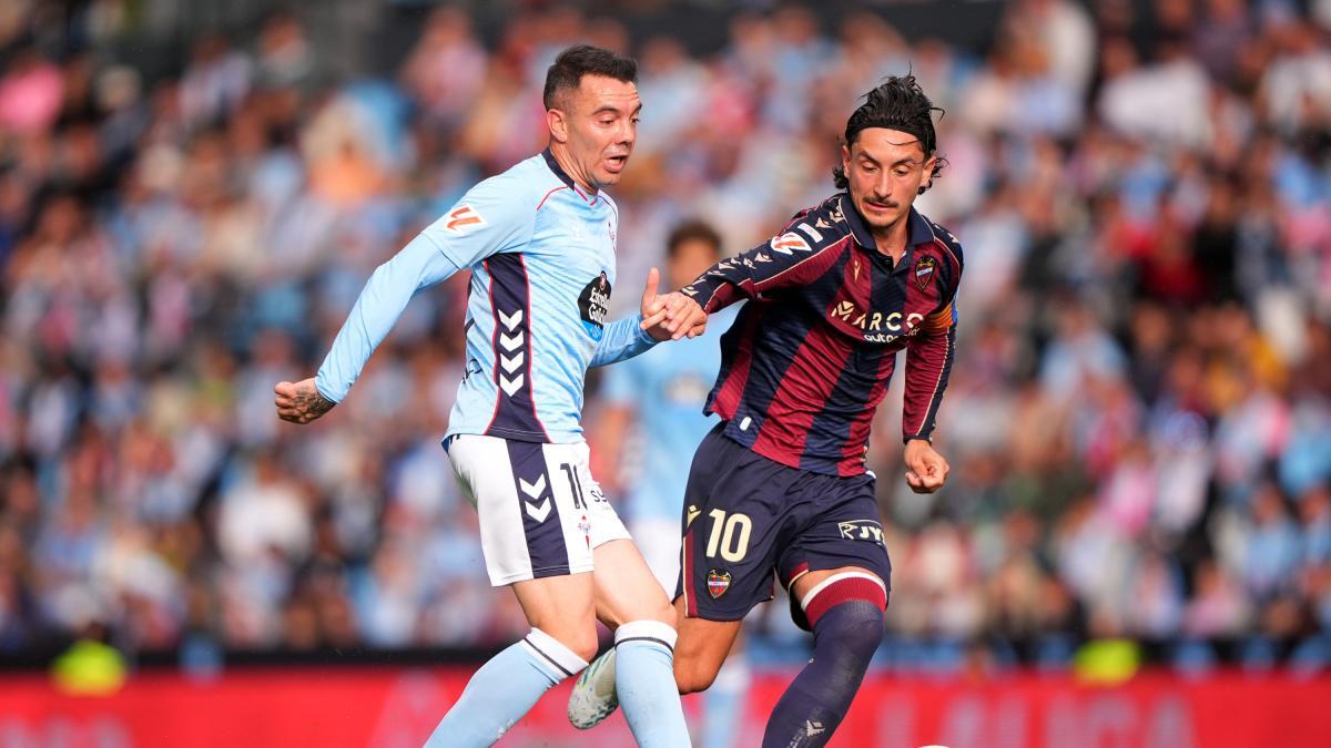 Two footballers compete for the ball during a football match, one wearing a white and light blue shirt and another in a red and dark blue uniform.