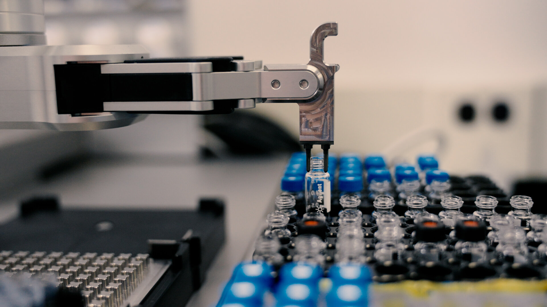 A precision robotic arm manipulates transparent and blue glass vials organised in a chemical reaction tray in what appears to be an automated laboratory.