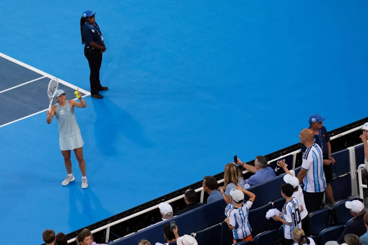 Una tenista en cancha azul sostiene una raqueta mientras observa hacia arriba, con una árbitra de pie en el fondo y espectadores en las gradas del lado derecho.