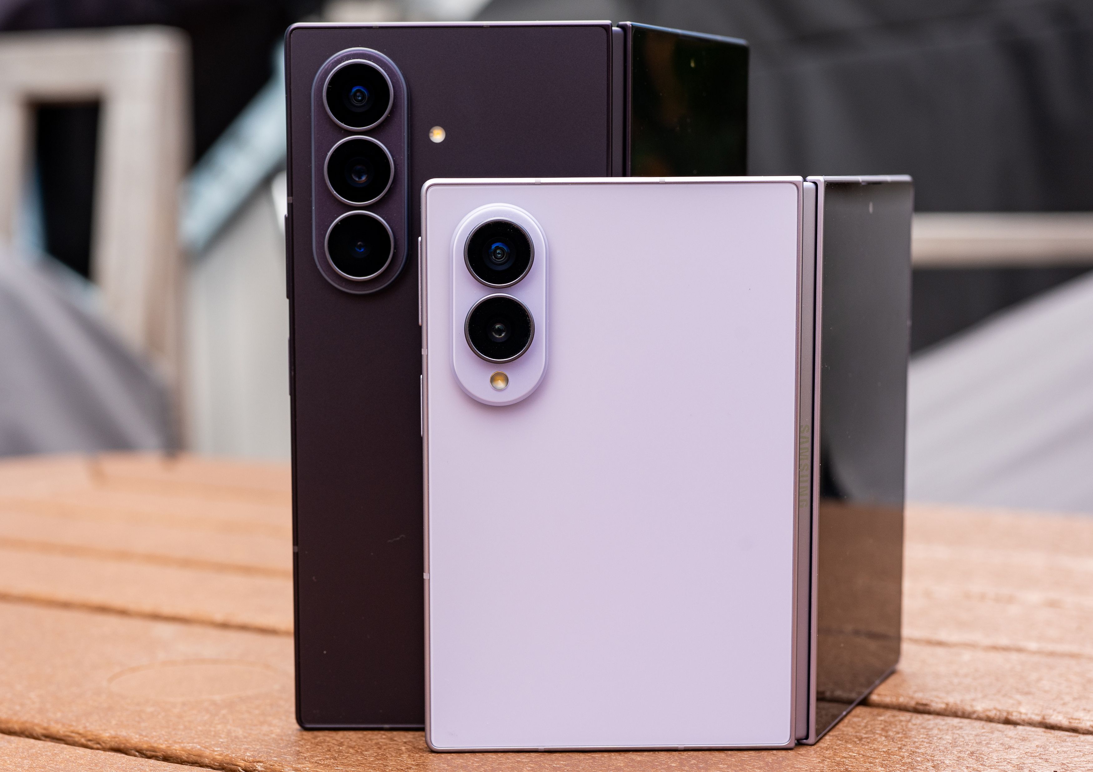 Two Samsung Galaxy Z Fold8 smartphones displayed side-by-side on a wooden surface, showing the back of the devices with their camera modules - the left phone in a dark burgundy color with three cameras, and the right phone in a light lavender color with two cameras.