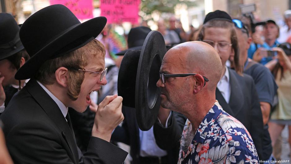 A man in a black suit and wide-brimmed hat faces off with a man wearing glasses and a floral shirt while holding a circular black object between them, surrounded by people on a busy Jerusalem street.