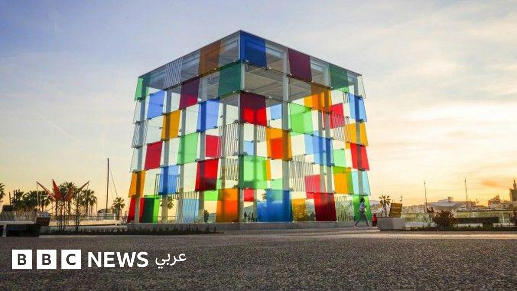 A cubic glass and steel building with bright coloured panels—red, blue, green, yellow, orange and white—arranged in an irregular geometric pattern in front of a maritime port at dusk.