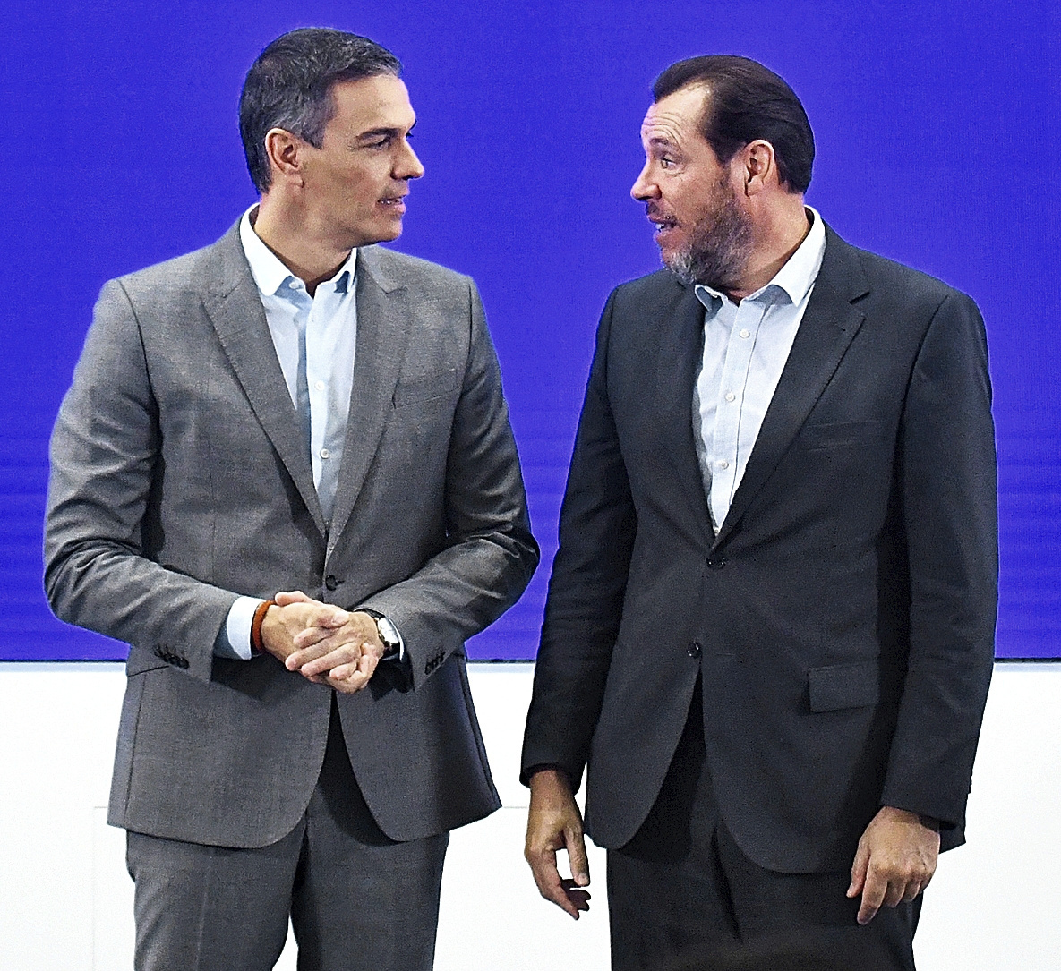 Two men in business suits engage in conversation against a blue background, with one man on the left in a gray suit and one on the right in a dark suit, both wearing light blue shirts.
