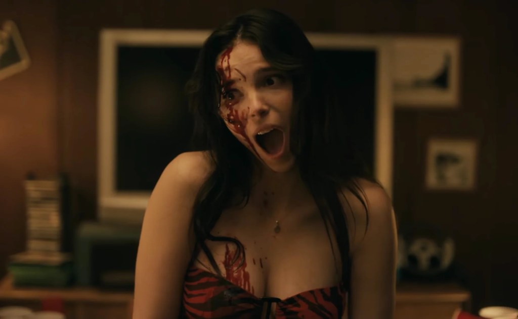 Una mujer con sangre en la cara y el cuerpo en un interior oscuro, con expresión de sorpresa.