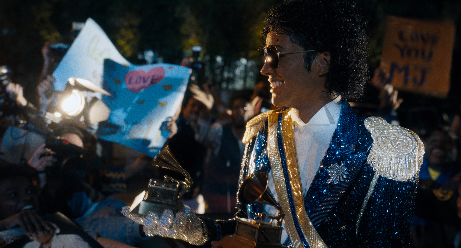A person in blue and gold sequined attire, sunglasses and a fedora hat stands in front of a crowd holding signs and phones at what appears to be a nighttime event.