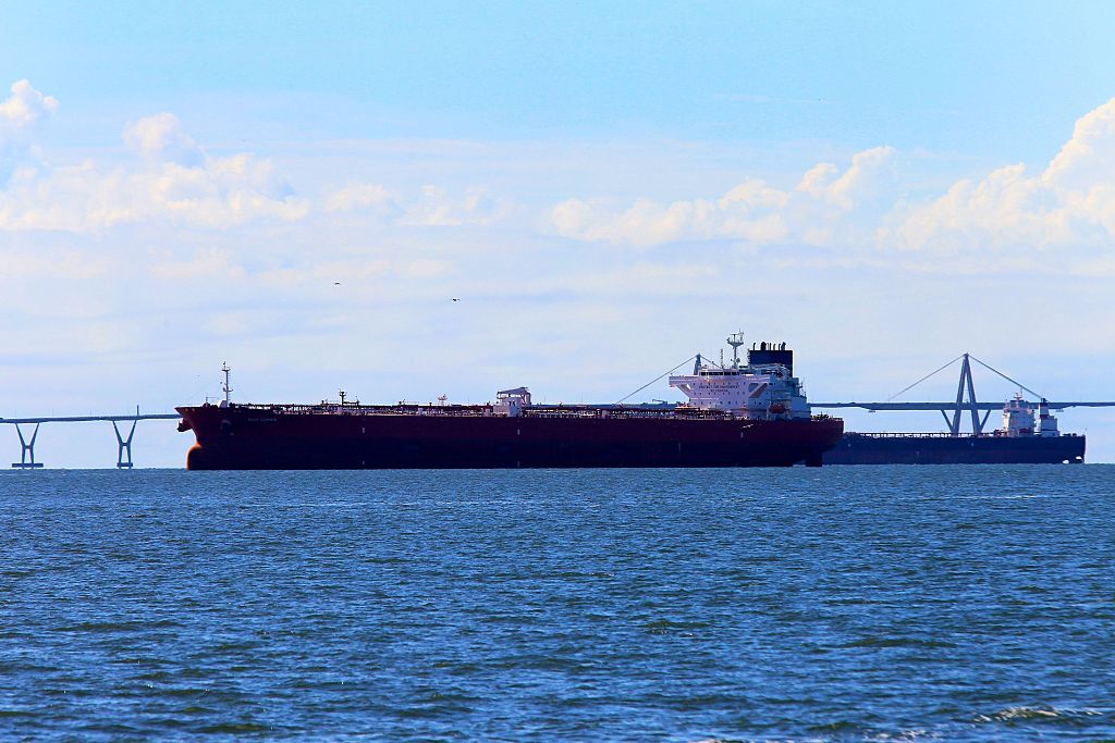 Un gran petrolero navega en aguas azules bajo un cielo despejado, con un puente colgante visible en el horizonte.