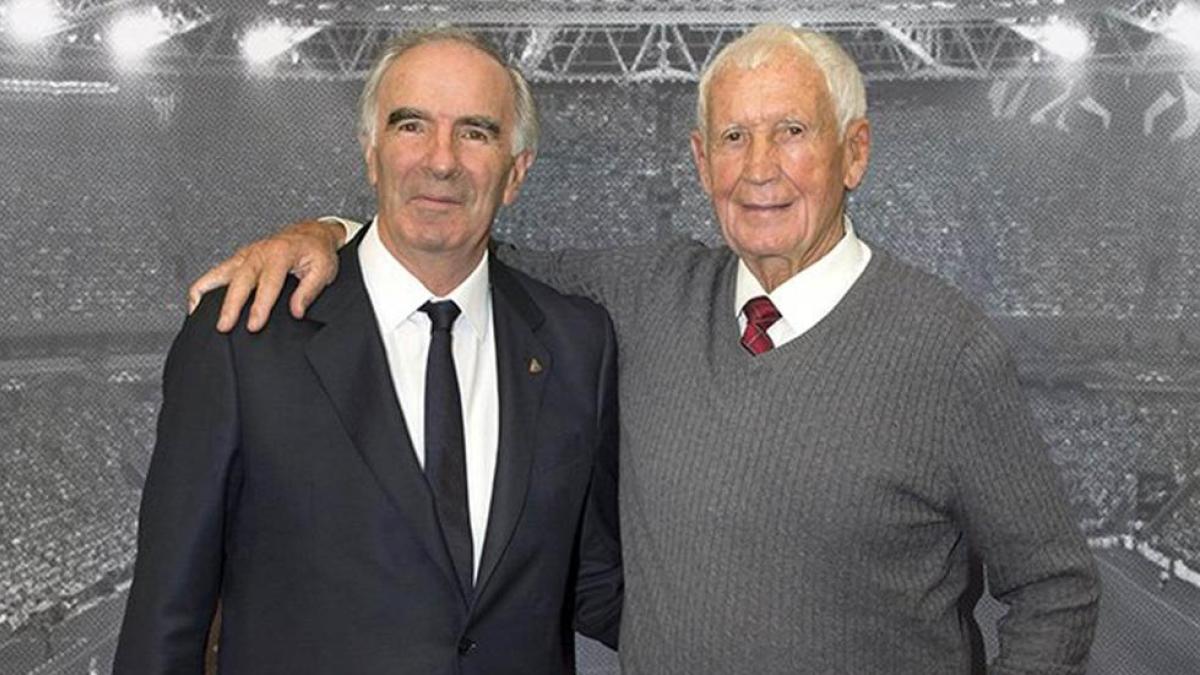 Two elderly men in formal attire stand together at what appears to be a stadium event, with their arms around each other's shoulders.