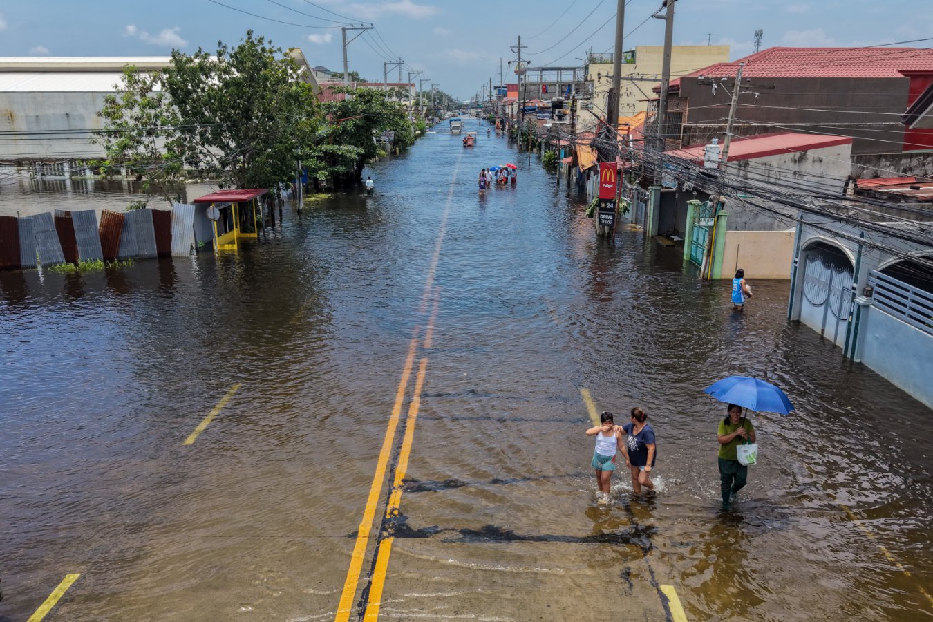 A completely flooded street in an urban area with multi-storey buildings on both sides, people walking through the water and a rainbow visible in the sky.