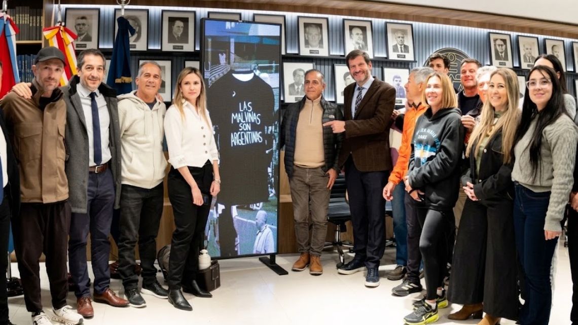 A group of people gather in a room with portraits on the walls while holding a black shirt with the text "The Malvinas are Argentine" and an image of a ship.