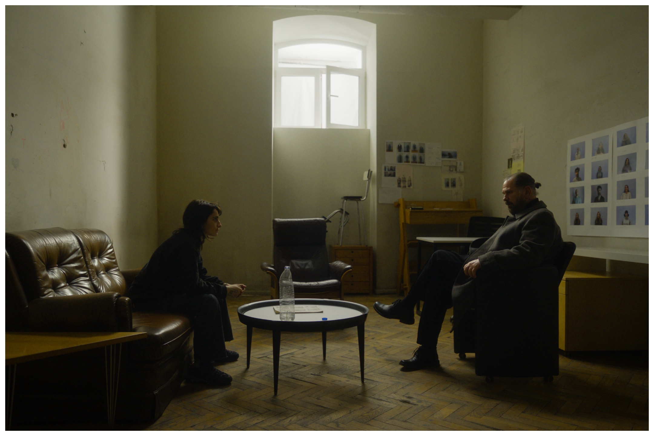Two men sit facing each other across a round table in a sparse, institutional room with pale walls, leather furniture, and windows showing bright daylight.