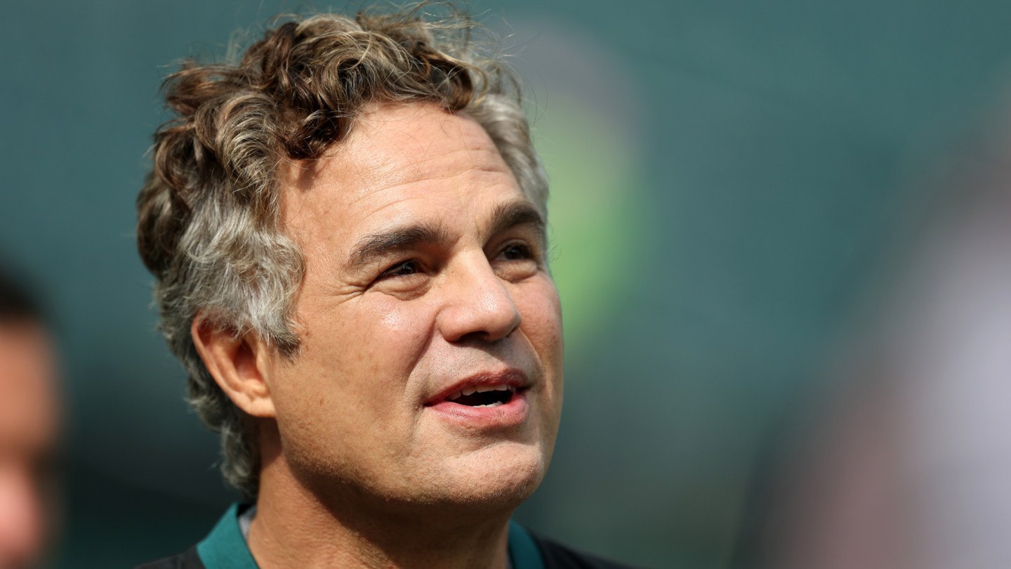 A middle-aged man with salt-and-pepper curly hair wearing a dark teal shirt speaks against a blurred teal background.