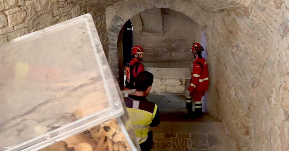 Firefighters in red protective gear and helmets work inside a stone corridor of a medieval building, with one person wearing a yellow safety vest visible in the foreground.