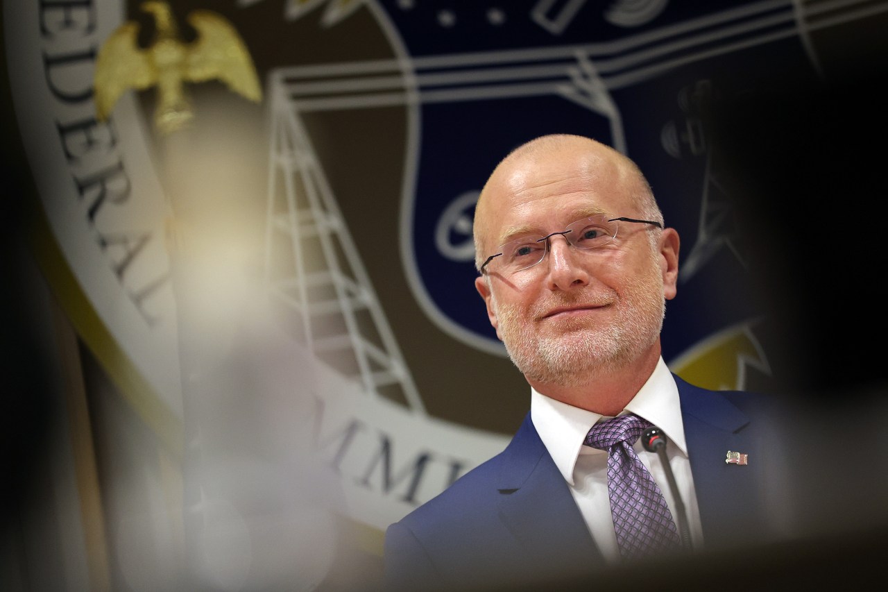 A bald man with glasses and a gray beard wears a dark blue suit and purple tie, standing in front of a blurred corporate or government backdrop with institutional imagery.