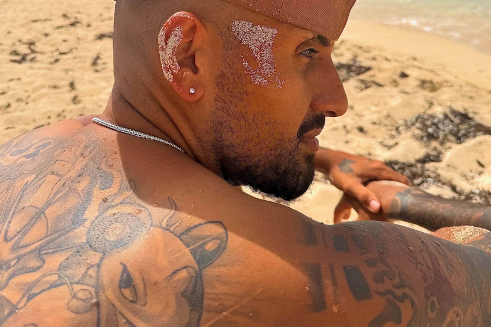 A man with tattoos on his arms and shoulder sits on a beach covered in sand, looking to the side in profile.