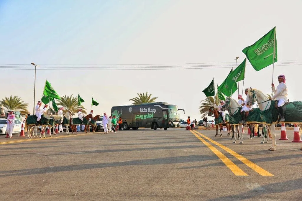 Parade on a wide road with people on camels holding large green Saudi flags, military vehicles, and spectators lining the sides under palm trees.