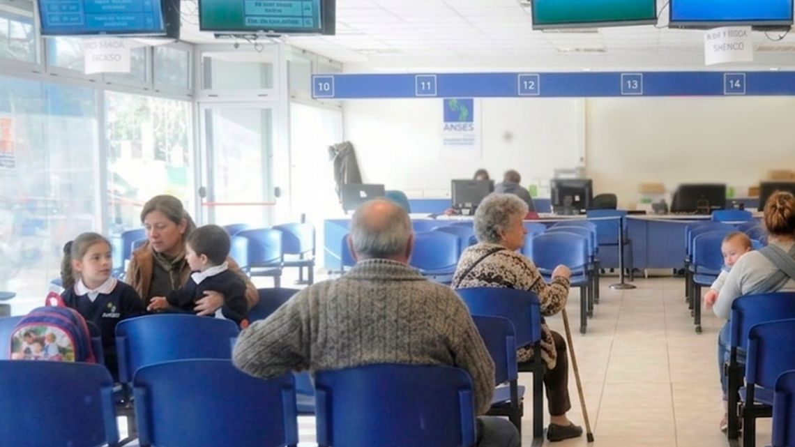 Personas de distintas edades esperan sentadas en sillas azules numeradas en una oficina de ANSES.