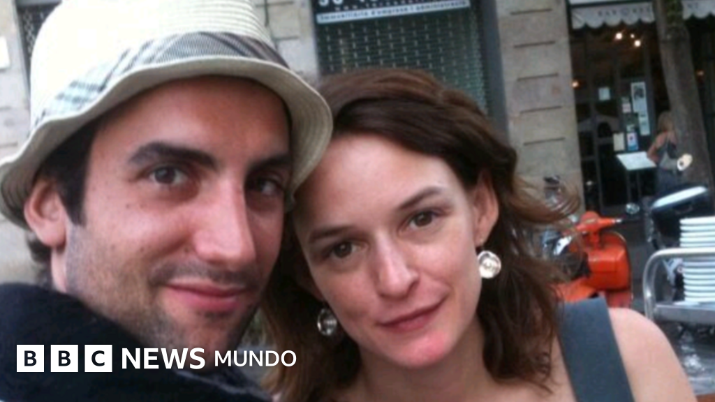 Un hombre y una mujer jóvenes sonríen juntos en una calle urbana, con la fachada de un edificio y un vehículo rojo visible al fondo.