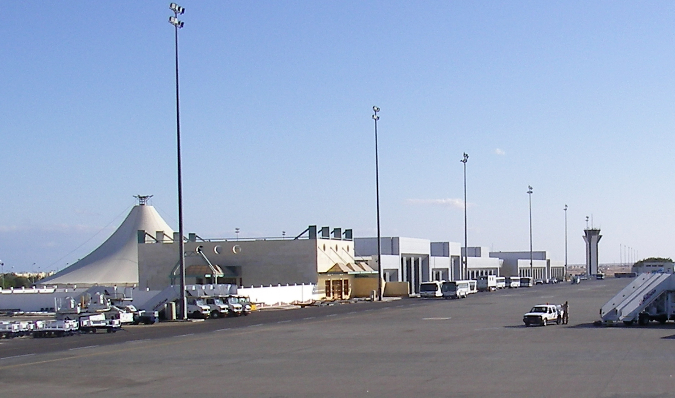 View of a modern airport terminal with a distinctive tent-like shaped roof, surrounded by tall lighting poles and support vehicles on the tarmac.