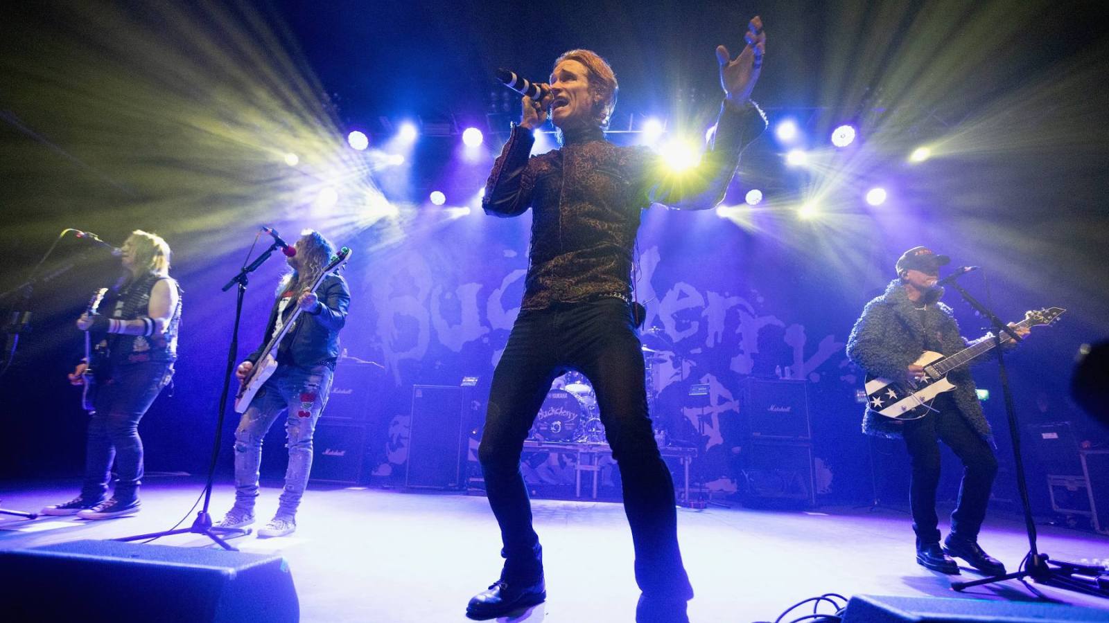 A rock band performing on stage with a lead singer in the center singing into a microphone, flanked by band members playing bass and guitar, with dramatic blue and white stage lighting in the background.