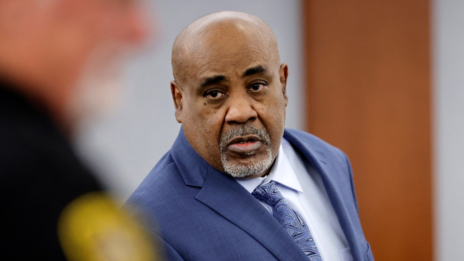 A man in a blue suit and patterned tie speaks while appearing in what looks like a courtroom setting.