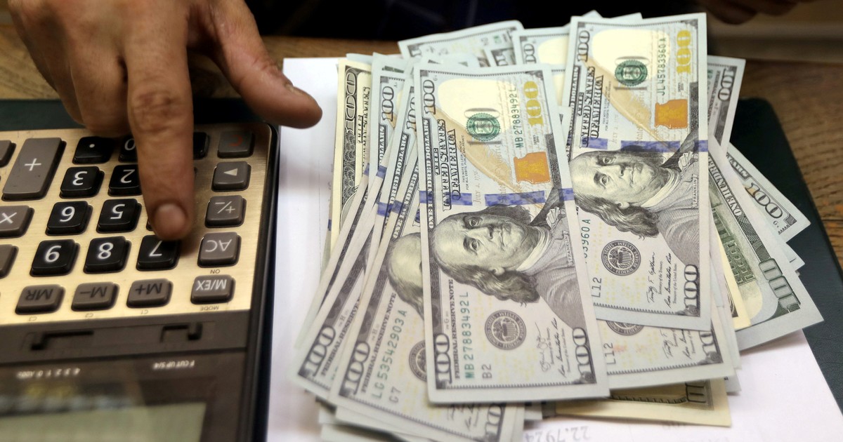 A hand holds several hundred-dollar US banknotes over a calculator.