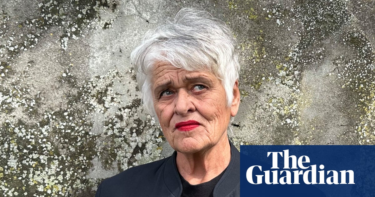 A person with short white hair and red lipstick wearing a black shirt stands against a textured gray wall with white lichen or moss.