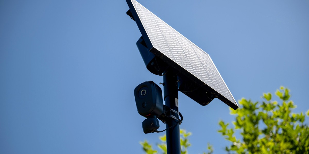 Una cámara de lectura de placas automáticas montada en un poste con un panel solar contra un cielo azul claro, con follaje verde visible en la parte inferior de la imagen.
