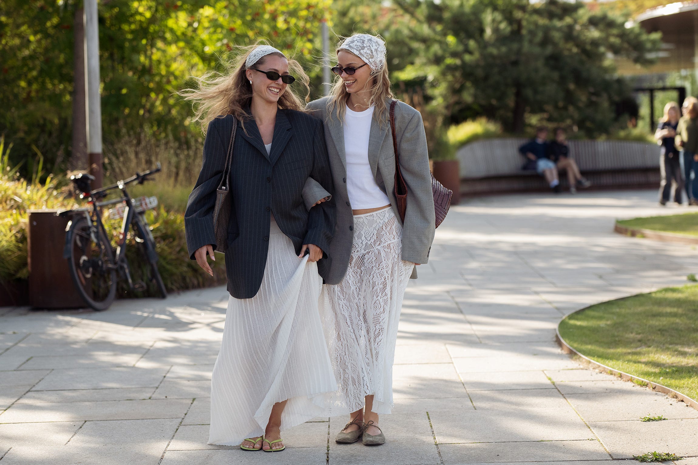 Two women walk smiling through a tree-lined promenade, wearing autumn clothing including oversized blazers, white pleated skirts and knitted accessories.