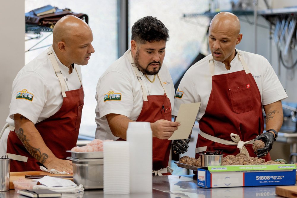 Three men in red aprons and white shirts work in a professional kitchen, preparing food while one holds a clipboard.