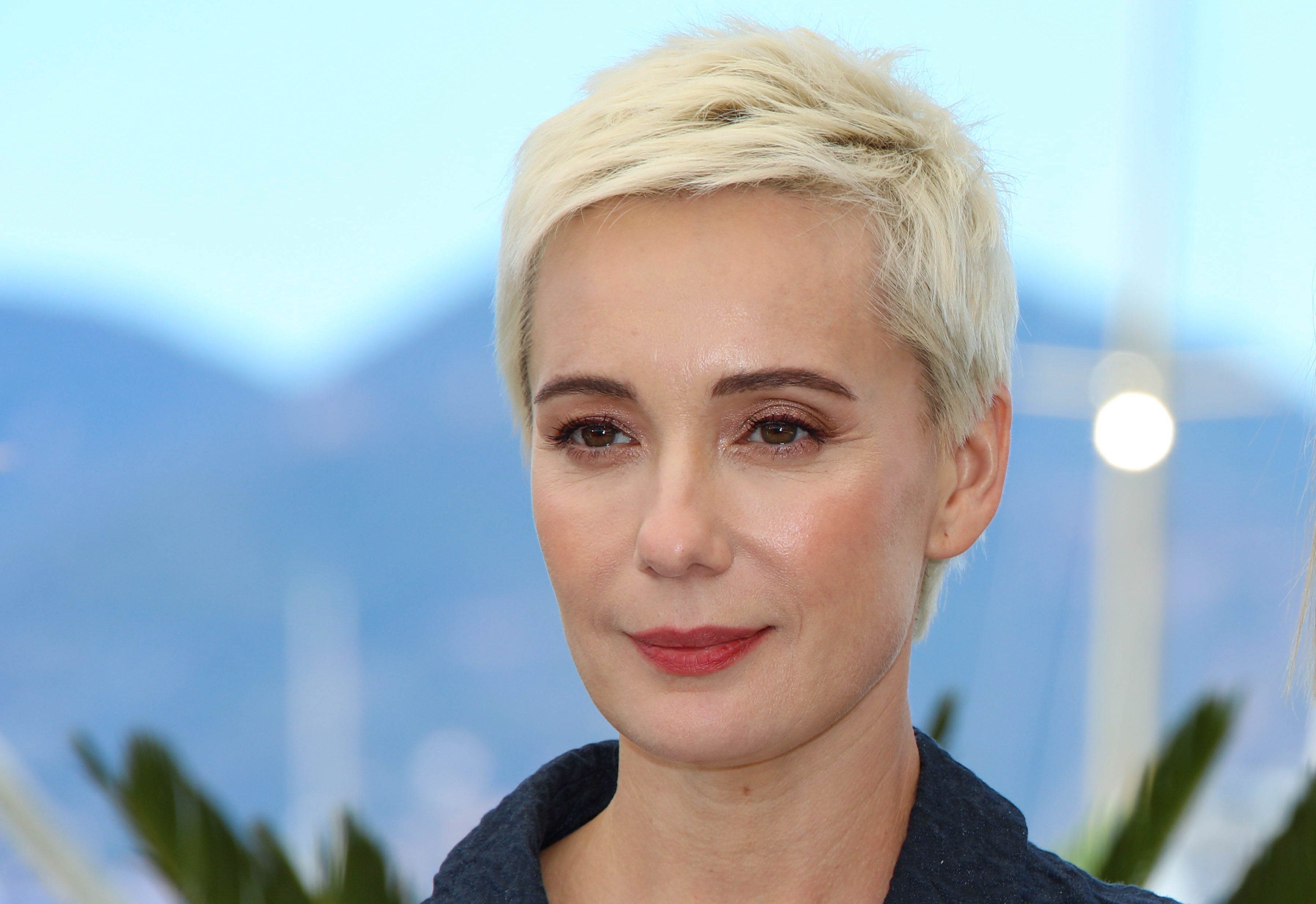 Portrait of a woman with short platinum blonde hair wearing dark clothing against a blurred blue cityscape background.