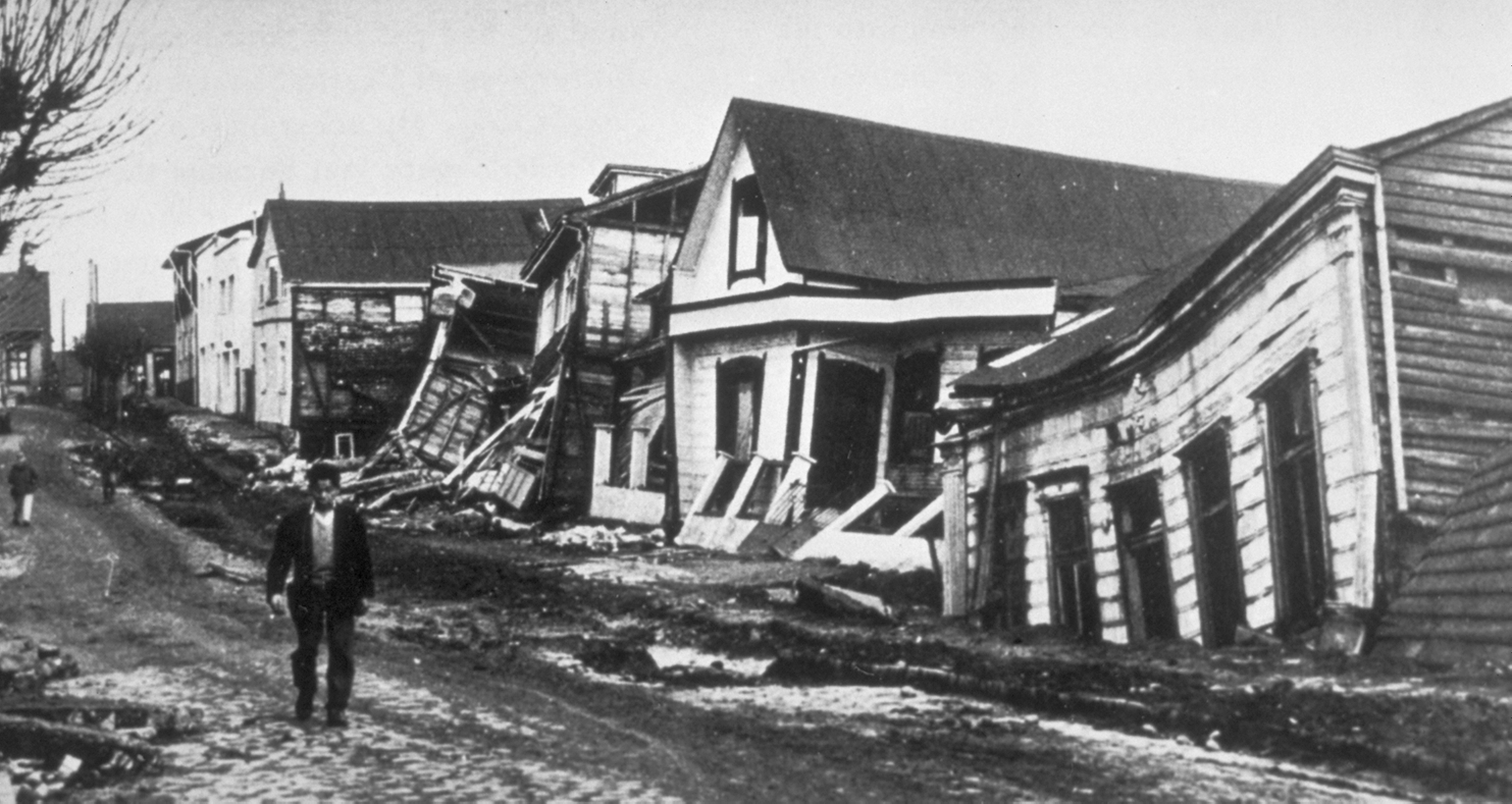 Fotografía en blanco y negro de casas de madera dañadas y parcialmente derrumbadas en una calle de tierra, con una persona de pie observando la devastación.