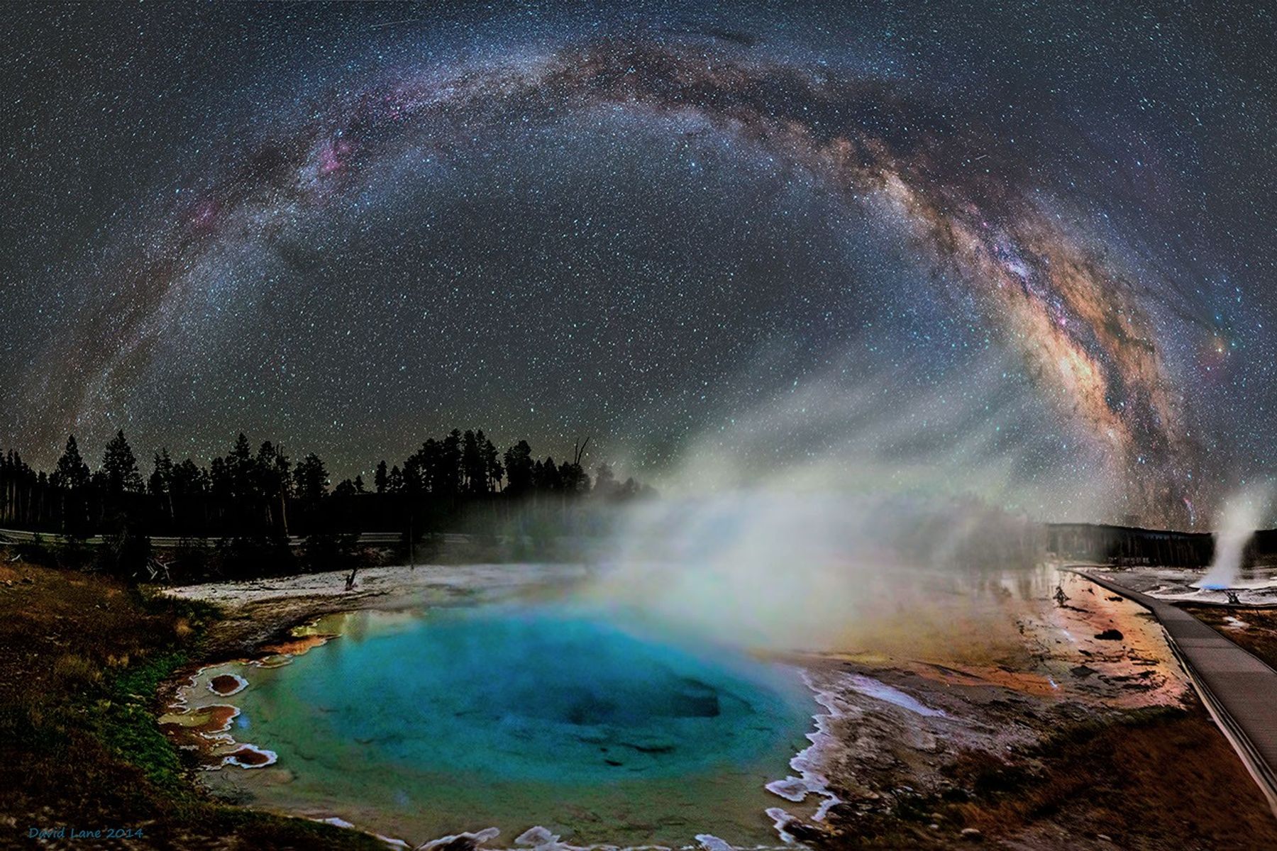 La Vía Láctea se arquea sobre un cielo estrellado oscuro, con un manantial termal azul brillante rodeado de vapor blanco en el primer plano y árboles de coníferas siluetados contra el horizonte.