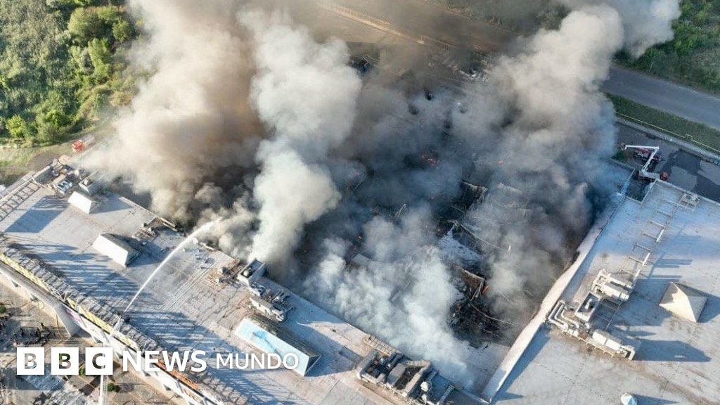 An aerial view of a commercial building engulfed in dense white and grey smoke, with flames visible among the damaged structures.