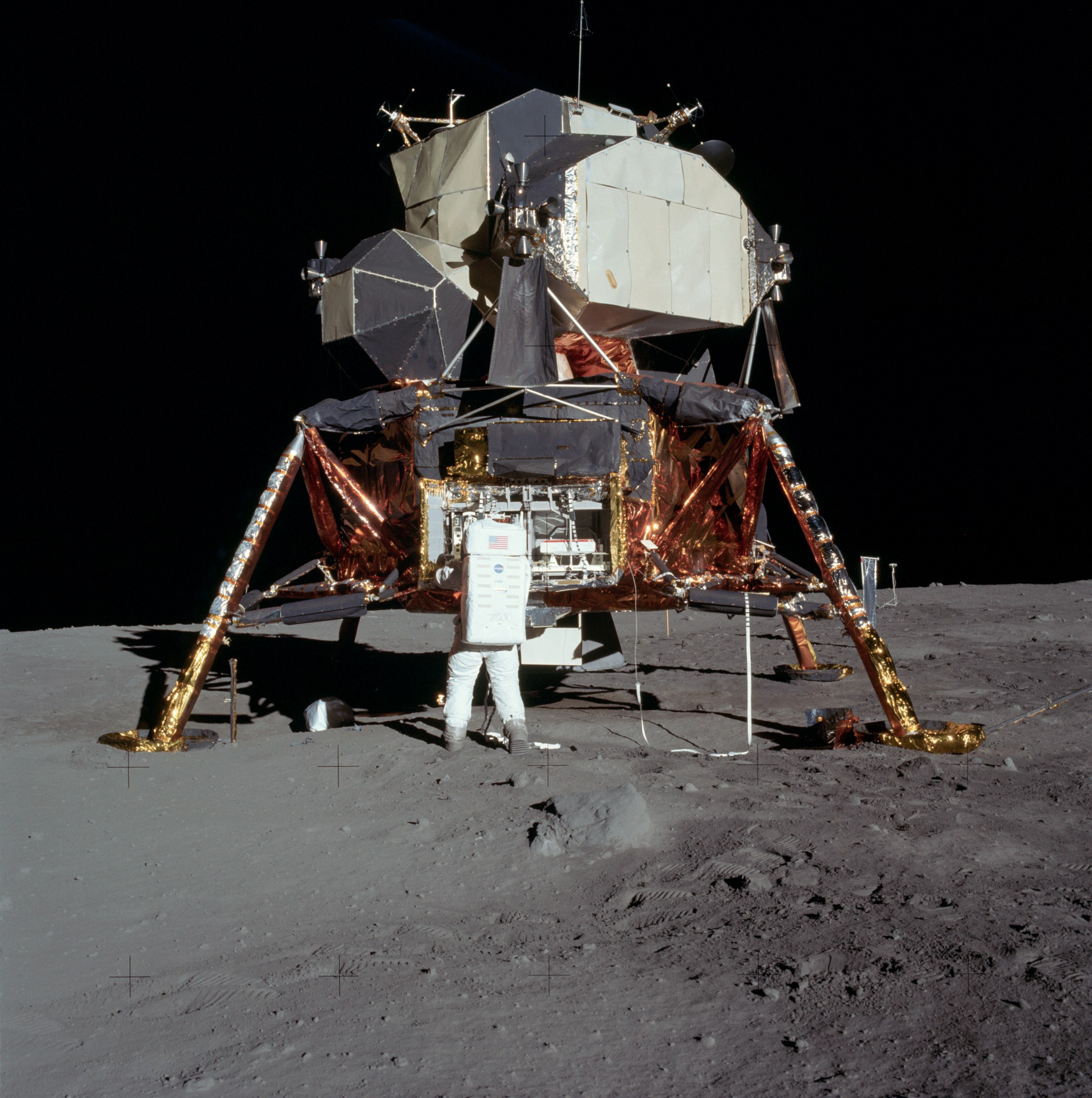 Un astronauta en traje espacial blanco parado en la superficie lunar junto a un módulo de aterrizaje lunar con paneles solares dorados y estructuras metálicas, contra un cielo negro.