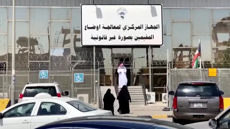 A building entrance with Arabic signage, parked vehicles, and two people in black clothing standing at the entry door.