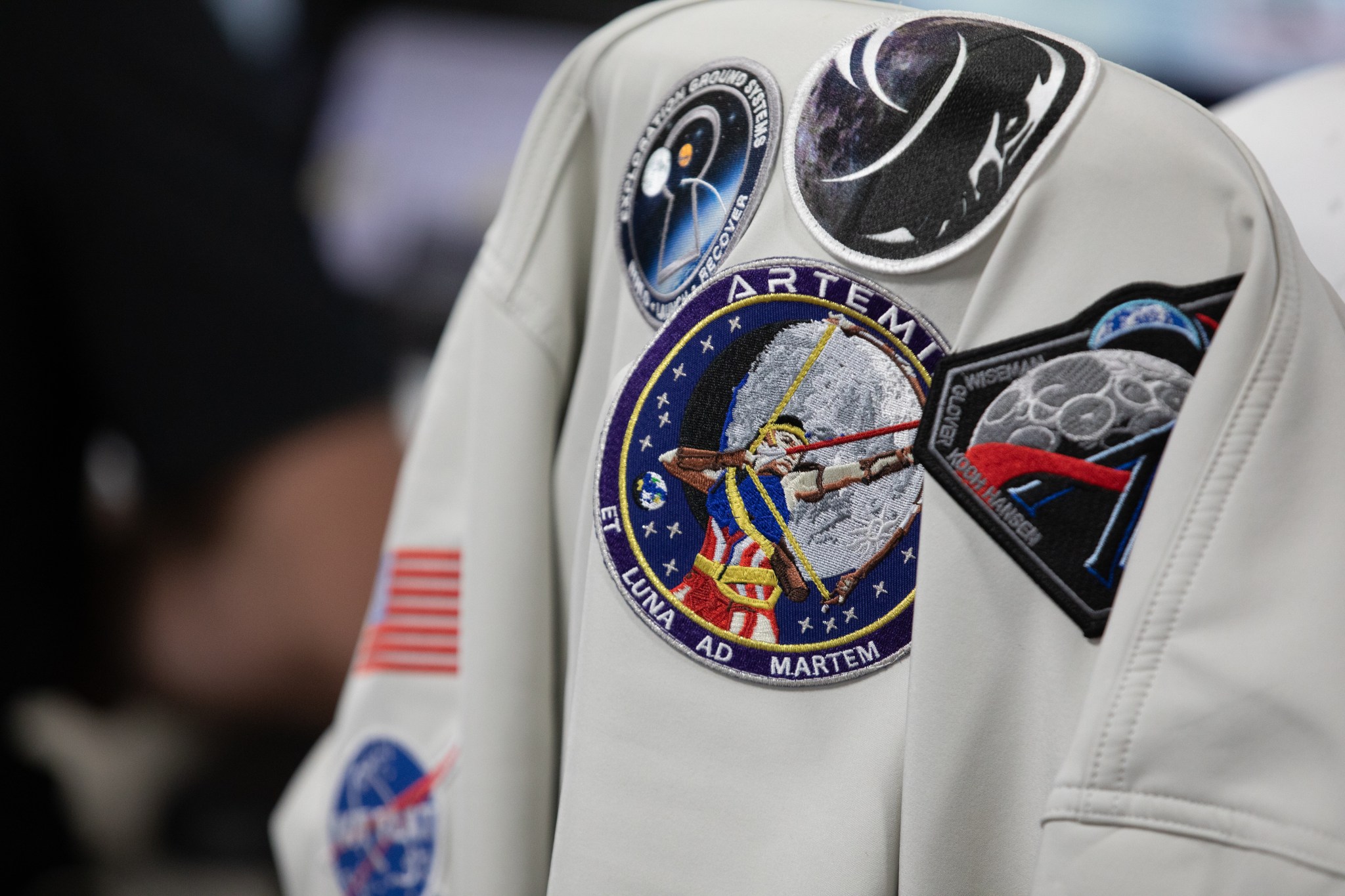 Una fotografía de cerca de un traje espacial blanco de la NASA con múltiples parches cosidos, incluyendo el emblema de la misión Artemis con la palabra "LUNA AD MARTEM" en el pecho.